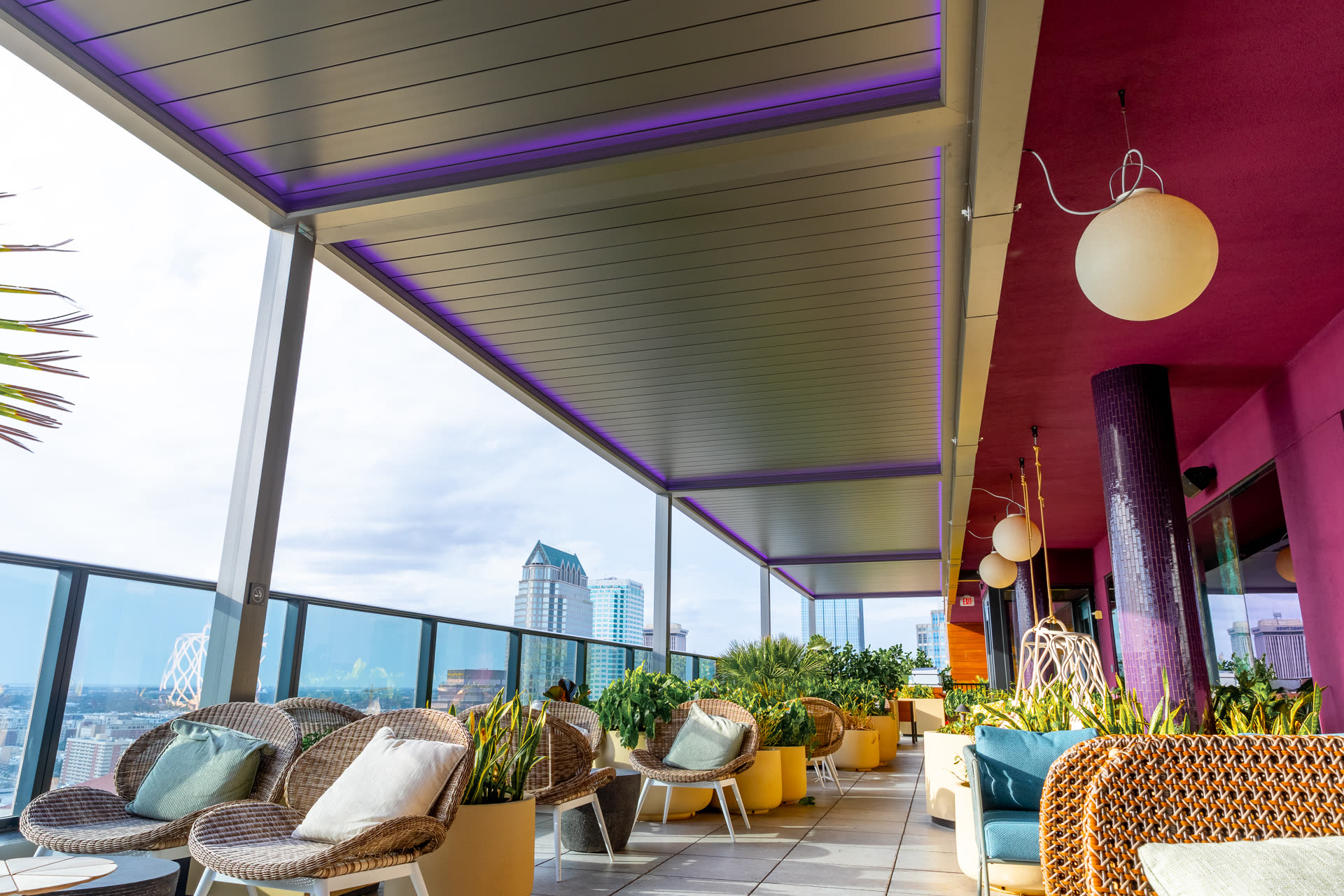 Rooftop terrace under a louvered aluminum pergola with violet LED strip lighting along the beams, wicker lounge seating, and a city skyline beyond. Manufacturer-network installation photo, not a CCSI project.