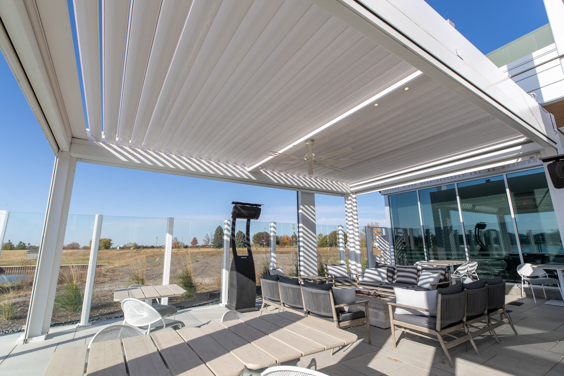 White Azenco R-BLADE pergola over a commercial lounge with partially open louvers, ceiling fan, patio heater, and integrated lighting. Manufacturer-network installation photo, not a CCSI project.