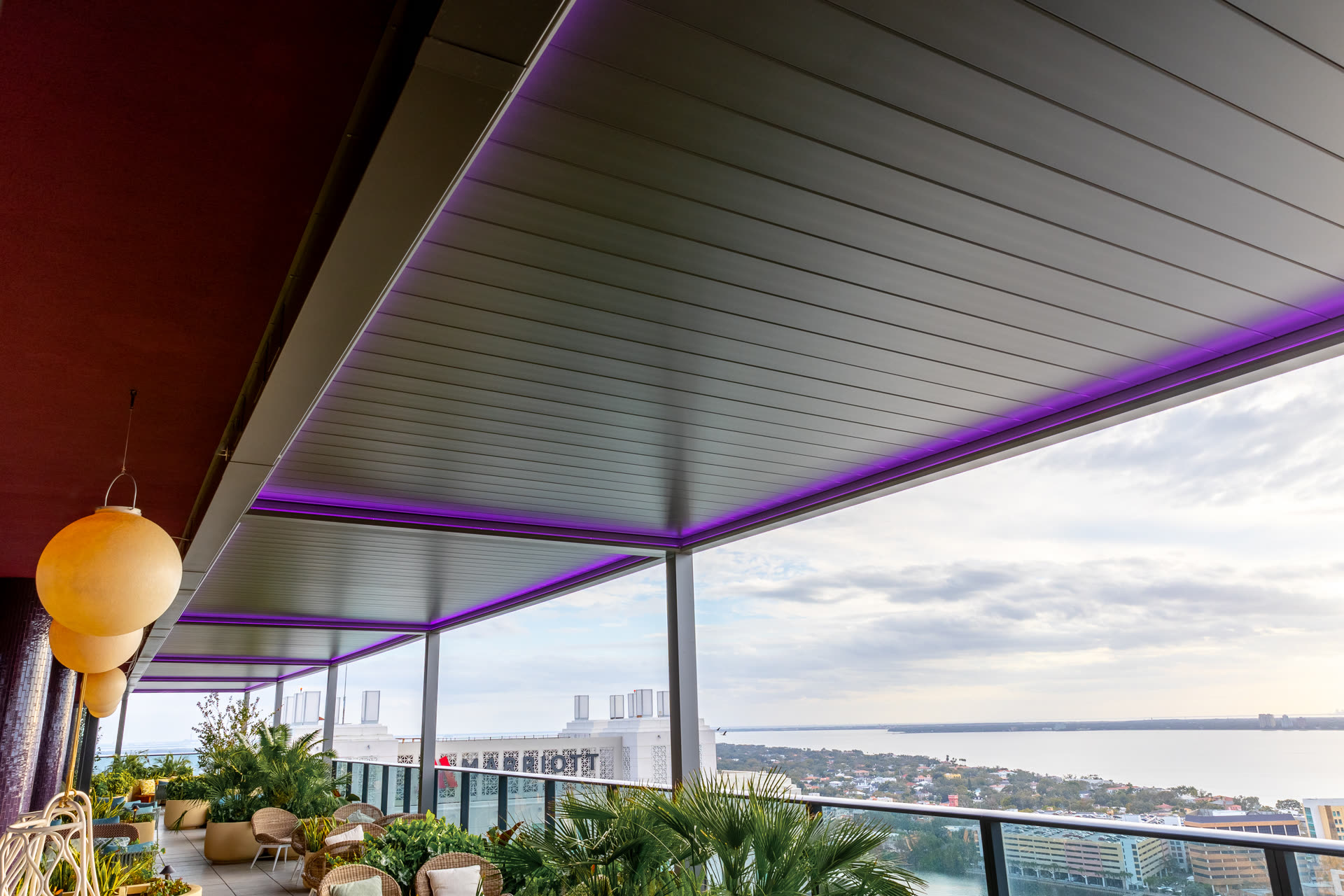 Louvered aluminum pergola running along a building facade on a rooftop terrace, with violet perimeter LED lighting and glass railings. Manufacturer-network installation photo, not a CCSI project.